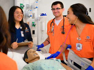 Four nursing students wearing orange and navy scrubs gather around a manikin in a hospital bed.