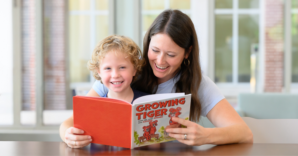 A child and an adult sit together at a table reading a brightly colored children's book titled Growing Tiger. The book features a cartoon tiger on the cover, and the pair are positioned in a bright indoor setting with large windows in the background.