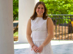 Graduation student marshal is pictured standing on a porch outside in a white dress.