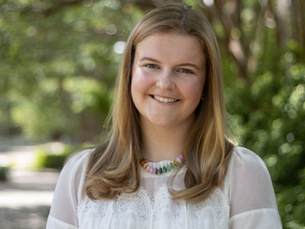 Whitney Shiver, a female nursing student wearing a white blouse and colorful necklace, stands in from of some greenery.