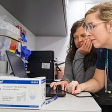 Two healthcare workers examine a computer screen in a clinical workspace, with medical supplies and a box of procedure masks in the foreground.