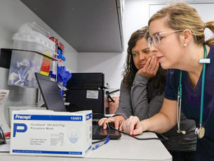 Two healthcare workers examine a computer screen in a clinical workspace, with medical supplies and a box of procedure masks in the foreground.