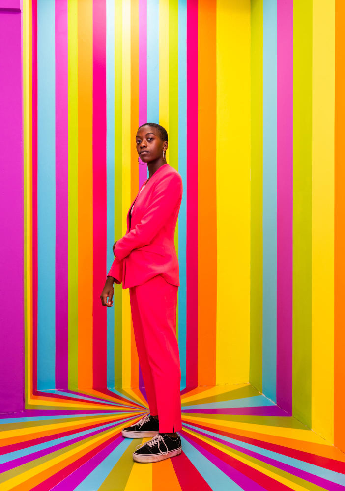 A confident individual in a striking neon pink suit stands against a vibrant background with vertical stripes in the colors of the rainbow. They have a composed expression, hands loosely by their sides, and are wearing black sneakers with white laces.