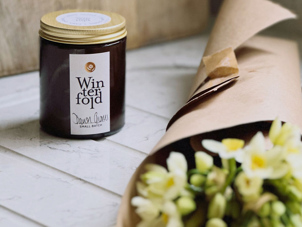 An candle in an amber jar sits on a marble counter with a bunch of lemon yellow narcissi
