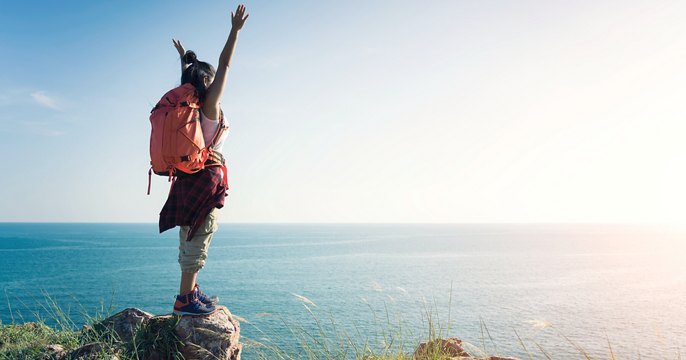 Girl with backpack hiking to ocean view with arms raised