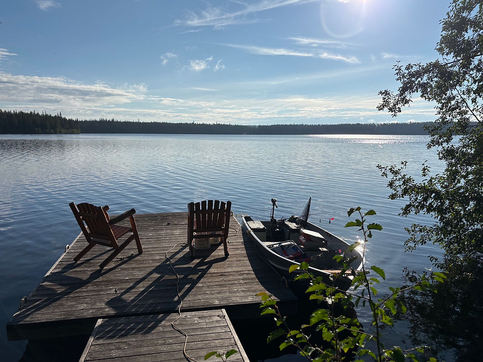 Bright, sunny morning lake view, showing two Adirondack chairs on dock casting long shadows in foreground with small, moored fishing boat at dock, and in the background a smooth lake with dark treeline and blue sky with few streaks of clouds; this scene of stillness contrasts with the busyness of the Fair just two days earlier