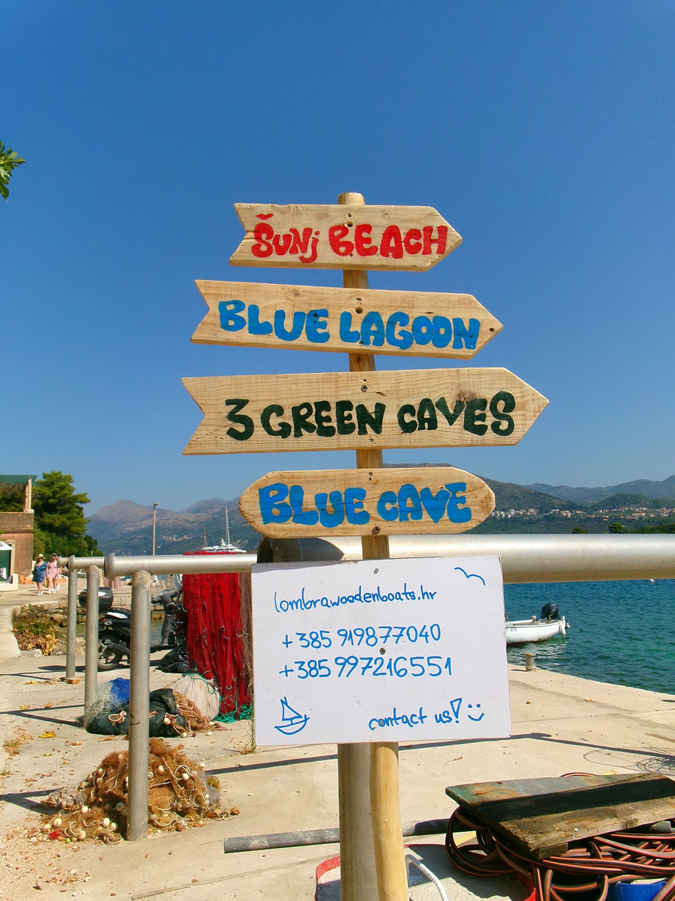 Wooden signpost with directions to beaches and caves in colorful text. Background shows a sunny harbor with boats and mountains.