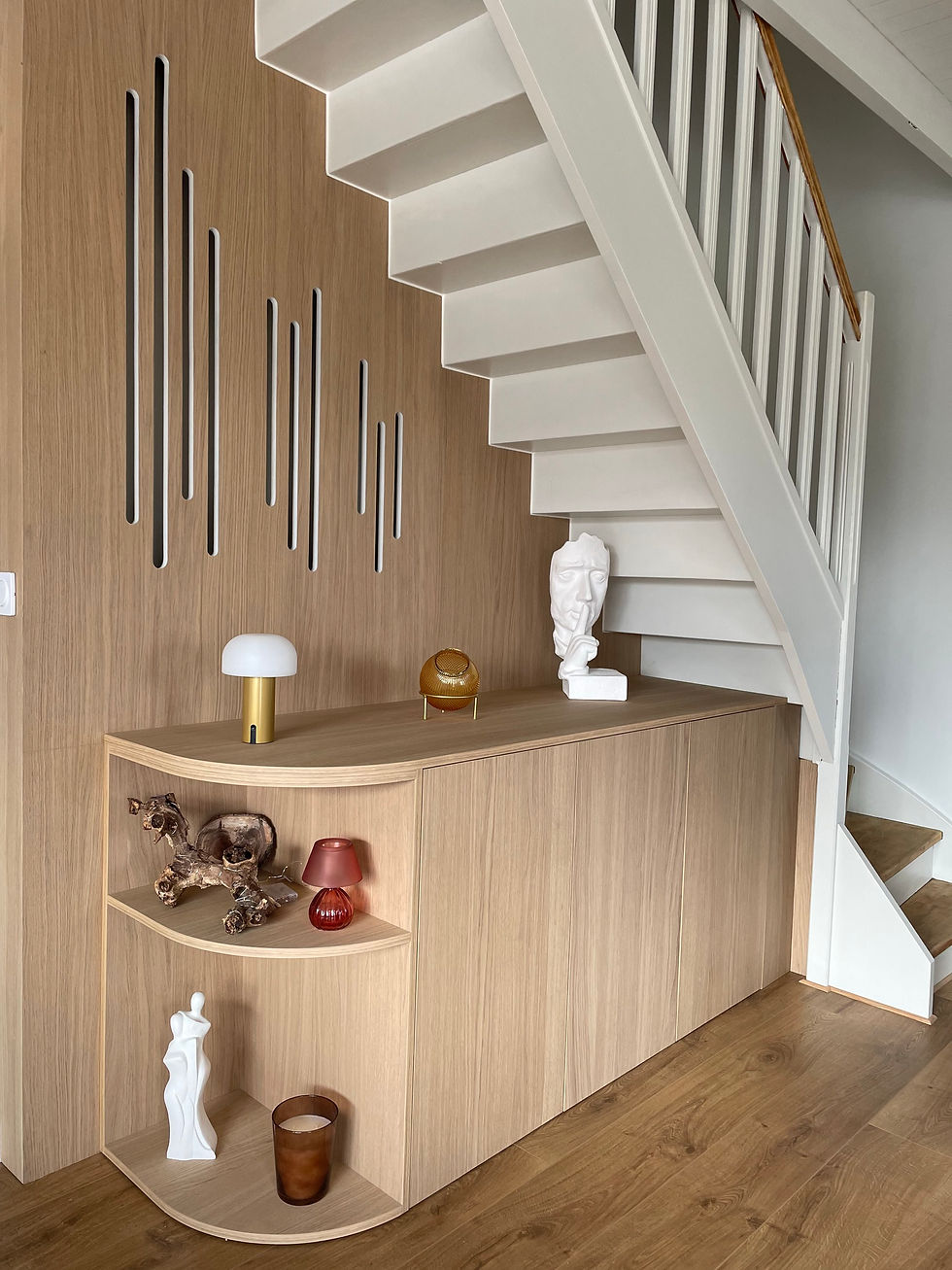 Meuble d’entrée sous escalier sur mesure réalisé par MAISONFABRIK à Saint-Omer, en placage chêne intégral, avec une finition arrondie élégante. L’aménagement comprend des caissons fermés avec ouverture push, ainsi qu’une partie ouverte en courbe avec deux niches pour exposer des objets décoratifs. Des découpes arrondies ont aussi été intégrées pour laisser circuler la chaleur des radiateurs, dans une logique de cache-radiateur aussi fonctionnelle qu’esthétique.