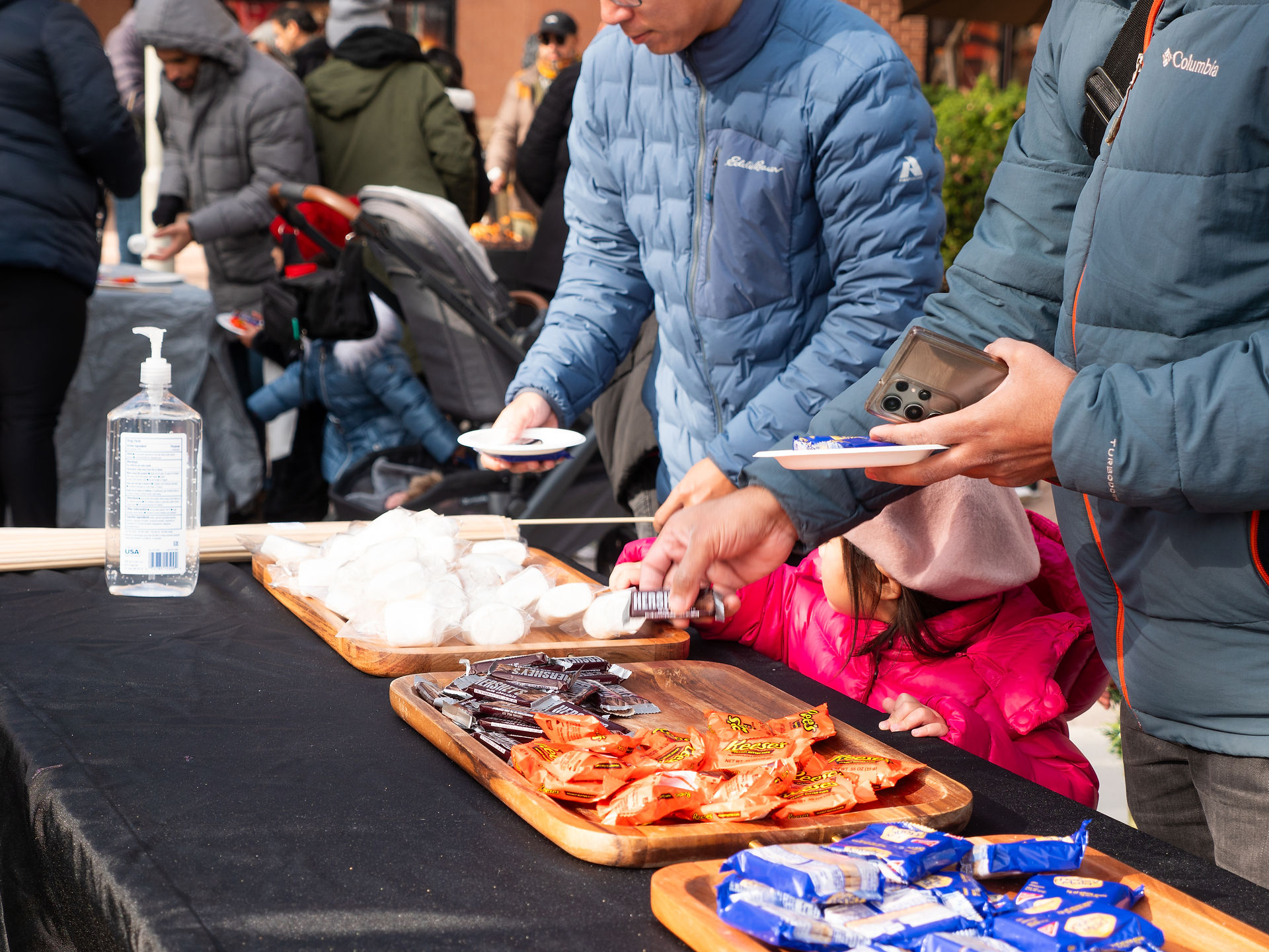 **S’mores Station at a Holiday Event** – Attendees gather around a table filled with marshmallows, chocolate, and graham crackers, preparing to make delicious s’mores. The cozy treat station adds warmth and sweetness to the festive outdoor gathering.