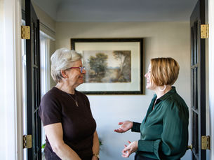Two women are smiling and having a conversation in a hallway. A landscape painting hangs on the wall behind them. Bright and friendly mood.