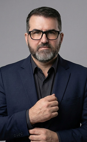 Foto do advogado Denis, homem de 50 anos com barba grisalha, óculos pretos e vestindo blazer azul marinho, camisa preta