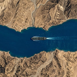 Aerial satellite view of an oil tanker navigating a narrow strait between desert mountain coastlines. Visual reference to the Strait of Hormuz, a chokepoint carrying one-fifth of global oil flows and central to India's energy security.