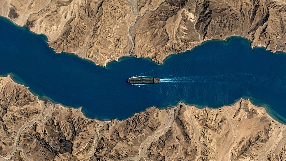 Aerial satellite view of an oil tanker navigating a narrow strait between desert mountain coastlines. Visual reference to the Strait of Hormuz, a chokepoint carrying one-fifth of global oil flows and central to India's energy security.