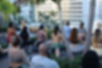 Group rooftop yoga session with instructor, participants wearing headphones, surrounded by palm trees and city buildings.