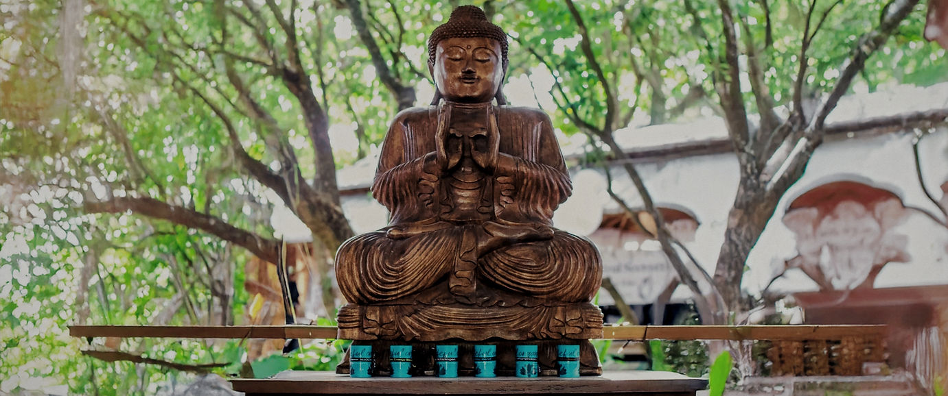 Wooden Buddha statue seated in a meditative pose on a platform outdoors, with small turquoise containers below and surrounded by lush green trees and rustic buildings