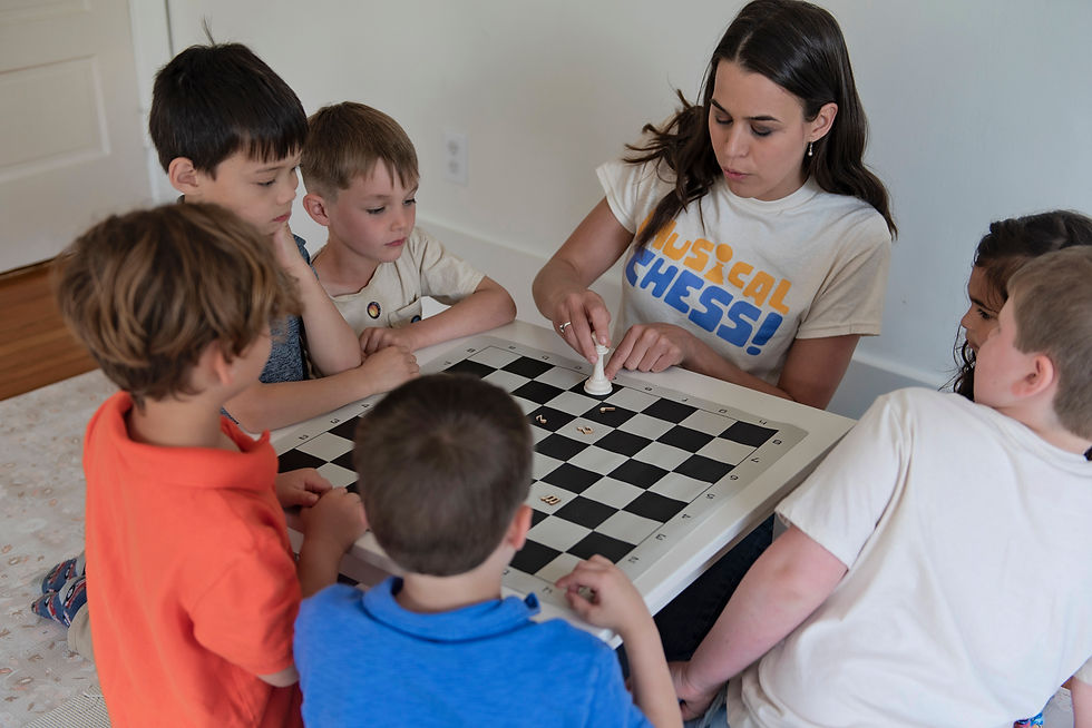 Founder Adrienne Tellier, in a "Musical Chess" shirt, teaches six kids around a chessboard. Children watch attentively.