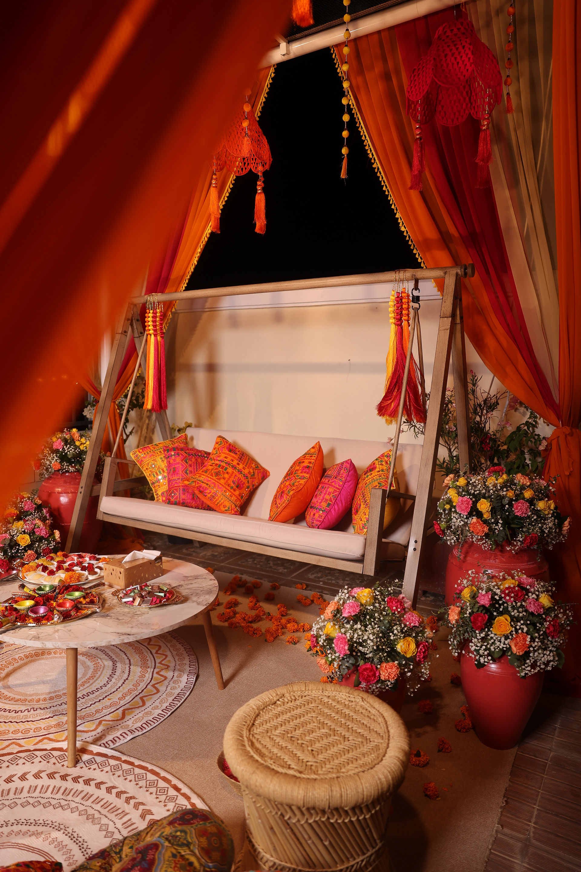 Vibrant Mehendi cabana with orange, pink, yellow drapes, marigold flowers, patterned cushions, and traditional Desi decor for bride and groom.