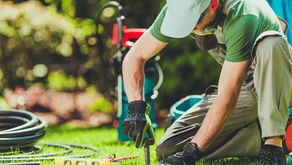 Gardener kneeling, installing a sprinkler on green grass. Wearing gloves and cap. Blurred greenery in the background. Tools nearby.