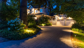 Illuminated suburban house at dusk with a paved driveway, blue hydrangeas, tall trees, and warm porch and garden lights.
