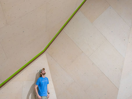 Operations manager looks up at a large wooden climbing wall with a green stripe. Bright setting, minimalist design.