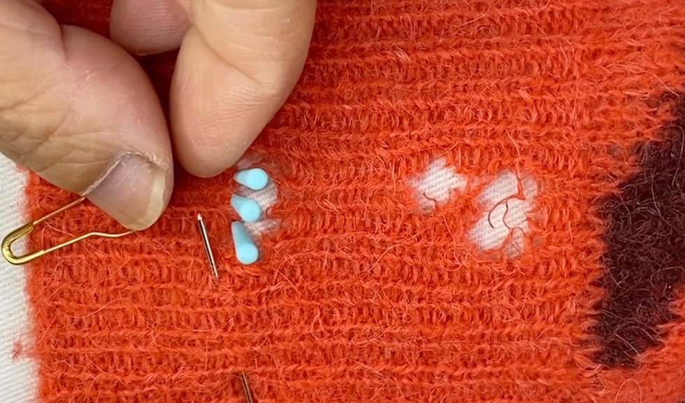 orangey red knit fabric with visible holes and hands of a Maker fixing the damage with pins