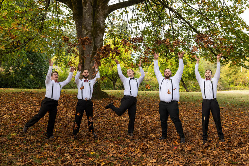 groom and four groomsmen in formal attire throw fall leaves in wedding shenanigan's