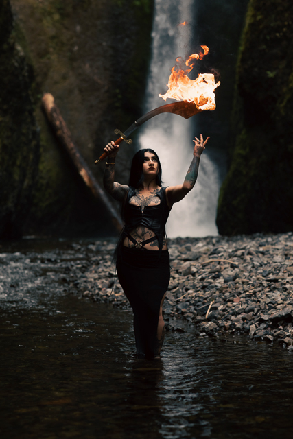 Golnar wielding a flaming sword in front of a majestic waterfall