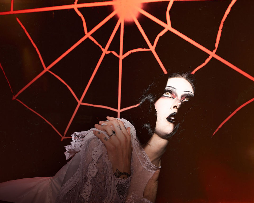 A model in white lace leans beneath the glowing spiderweb with a hand on her shoulder.