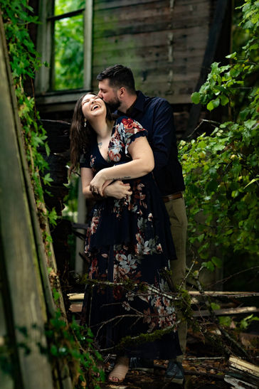 Snuggling couple laughing in front of abandoned overgrown building