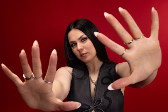 Close-up of musician Jessica Rook facing the camera against a red backdrop, both open hands reaching toward the lens.