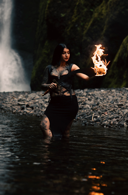 Golnar wielding a flaming sword in front of a majestic waterfall