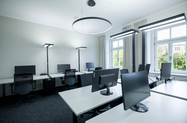 Fix Membership Bereich im 1. Obergeschoss von Sissi Works Coworking Space Wien. Höhenvertsellbare Schreibtische, Arbeitslampen, Bürostühle, dunkler Teppichboden, Blick aus dem Fenster in den grünen Garten
