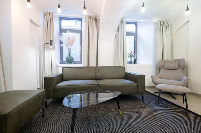 COACHING ROOM im Untergeschoss. grüne Couch, schwarze Stehlampe, dunkler Tisch, beige Vorhänge und Blick aus den Fenstern.