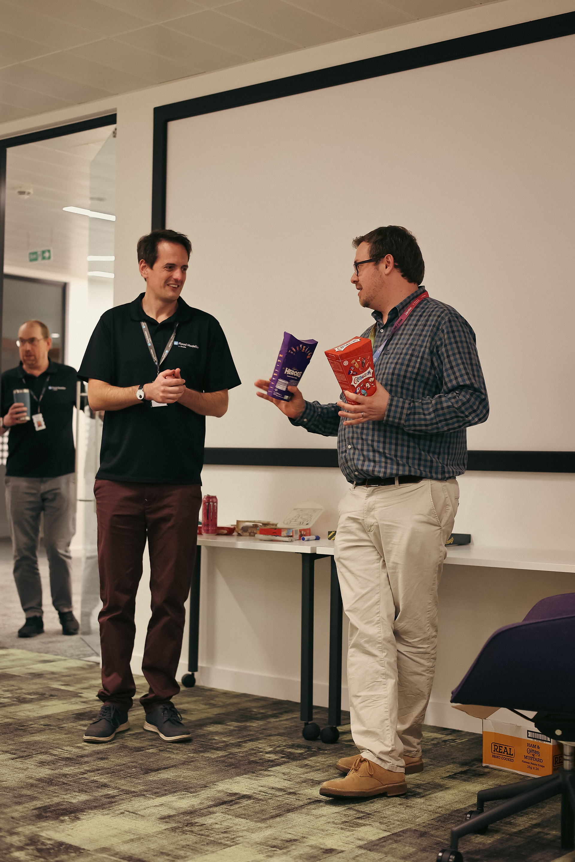 two people stood up with one of them holding two boxes of chocolates. there's a whiteboard in the background
