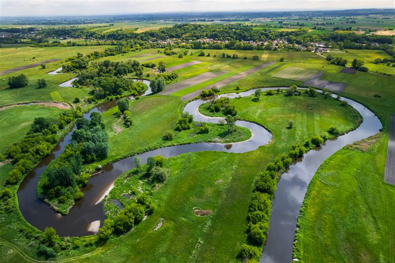 Aerial view of a winding river through lush green fields and patches of farmland, with a distant village under a cloudy sky.