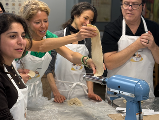 Four people  at an AI startup company team building event in aprons making ramen noodles with a blue machine. One holds dough; others watch inquisitively. Bright kitchen setting.
