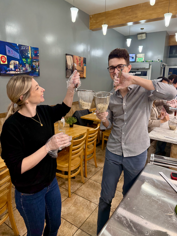 A happy couple at a ramen cooking class in San Francisco, playfully holding up baskets of freshly made noodles. This image captures the fun and interactive experience of a unique date night.