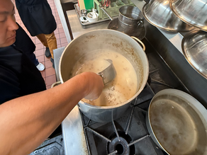 Person stirring creamy tonkotsu in a large pot on a stove, with metal pots nearby. Kitchen setting with tiled floor. Steam rising.