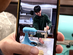 Person making ramen noodles with a blue machine, viewed through smartphone camera. Kitchen setting, stainless steel shelves in background, focused mood.