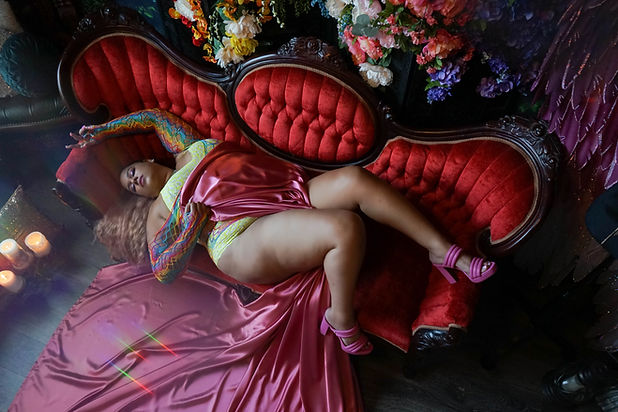 A plus size black female laying seductively on a red Victorian couch covered in a silk pink sheet.