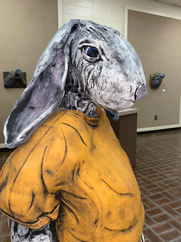 Rabbit sculpture wearing blue pants and orange shirt in a gallery space.
