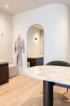 Modern, medical consulting room interior with desk in foreground, medical treatment bed and mirror beyond.