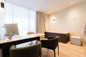 Modern plastic surgeons consult room with stone desk, dark timber accents and custom treatment bed beyond.