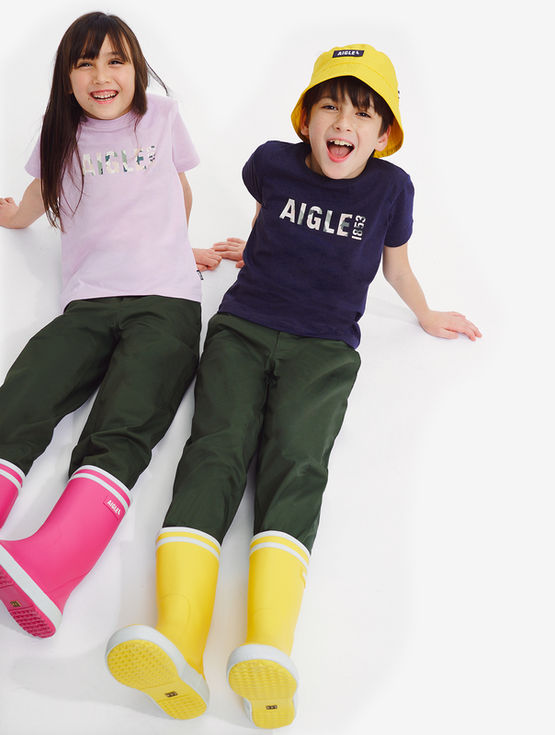 Two happy children wearing "AIGLE" shirt, green pants, and colorful boots.