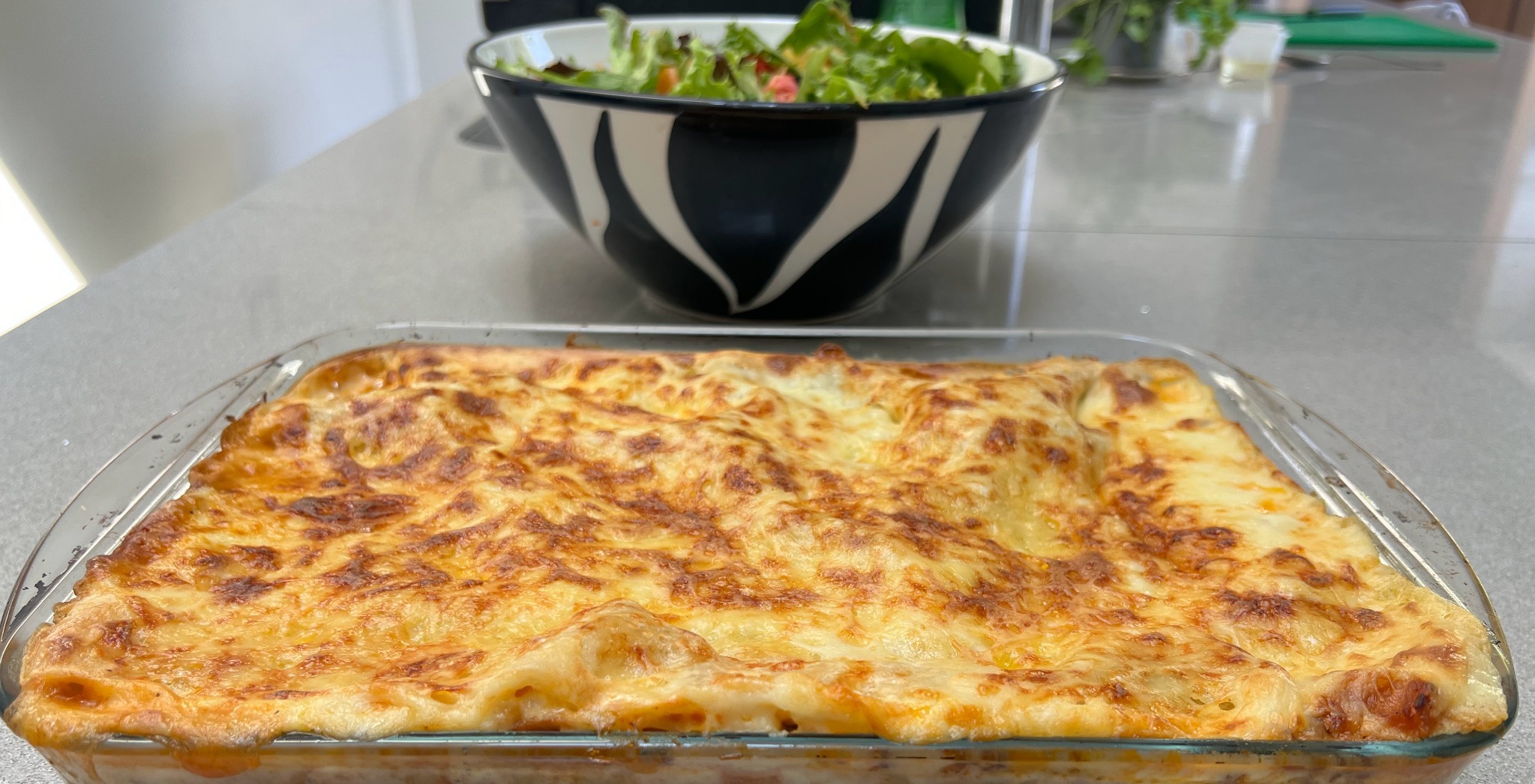 Vegetarian lasagne baked in a glass sharing dish, with a fresh salad behind it, by Noba Kitchen.