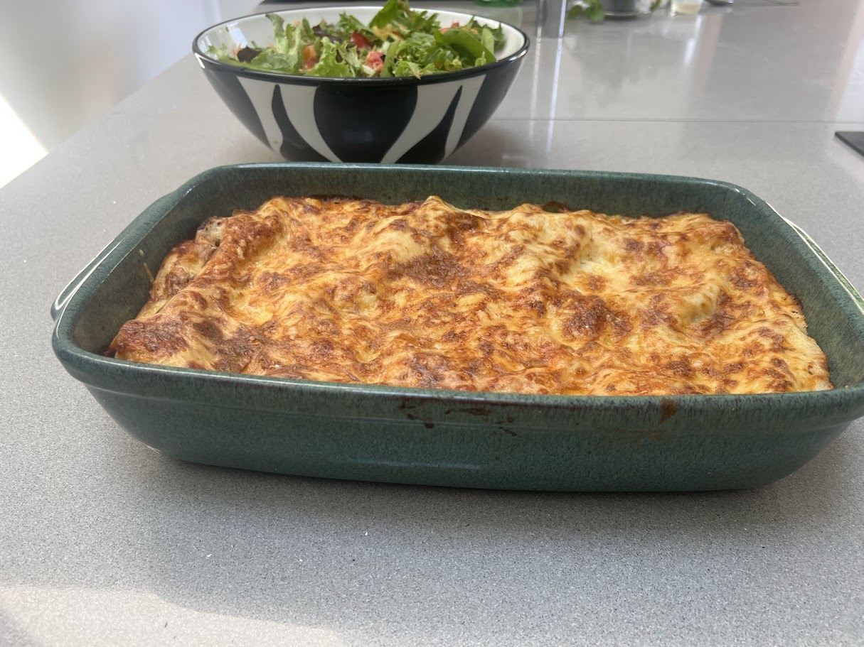 Beef lasagne baked in a green sharing dish, with a fresh salad behind it, by Noba Kitchen.