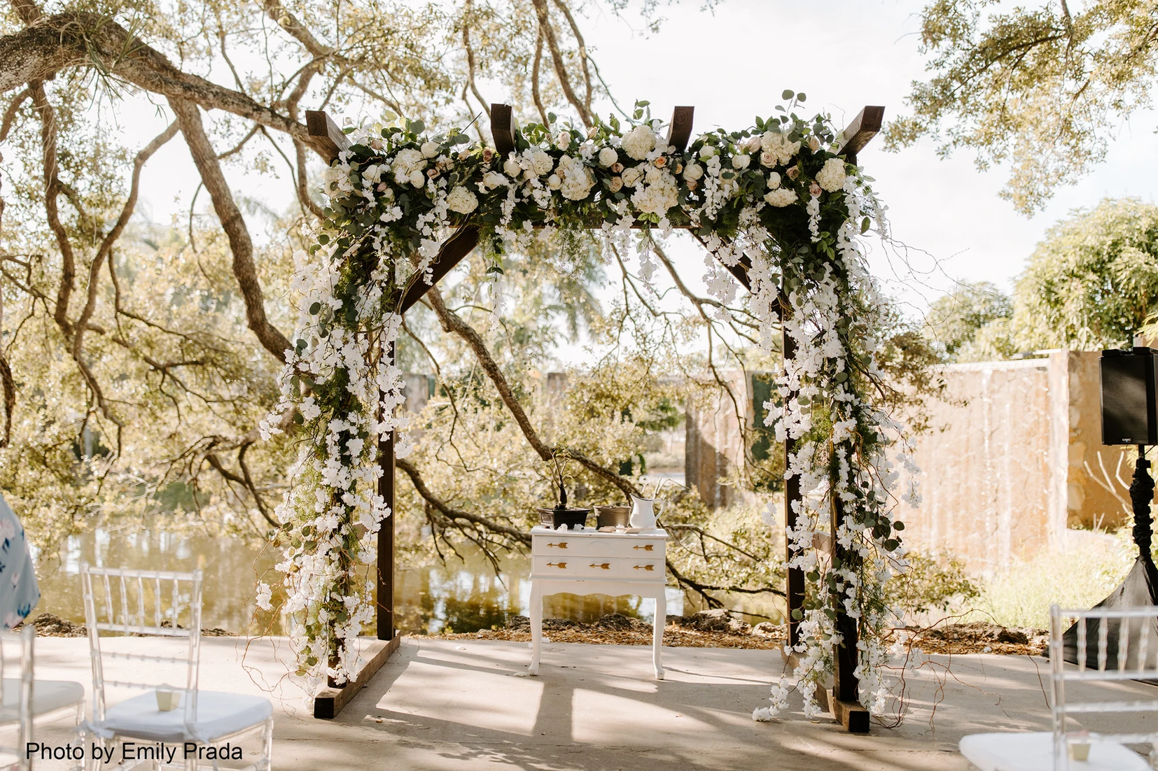 Wedding arch covered in white flowers with sun rays shining down between the trees and onto the arch