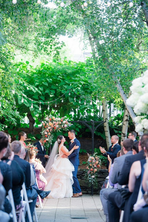 Newlyweds embrace surrounded by green foliage during outdoor garden ceremony.