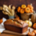 Loaf of banana bread on a wooden board, surrounded by pumpkins and bananas. Warm, rustic setting with autumn colors.