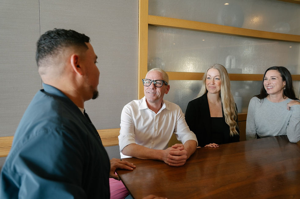 Four coworkers smile in a modern office meeting room around a wooden table, listening and chatting.